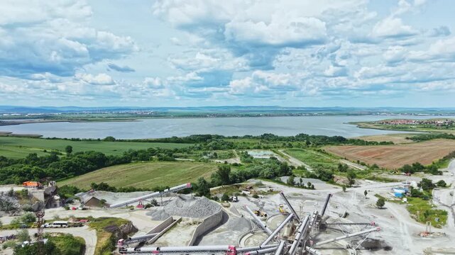 Aerial industrial quarry with exposed rock, heavy machinery moving gravel, stockpiles and access roads, adjacent wetlands and farmland, survey perspective