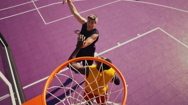 Two diverse basketball players compete on a vibrant purple outdoor court, one soaring for a dunk from above, capturing athletic training, energy, and dynamic urban streetball action