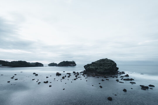 Long exposure seascape of rocky formations and scattered boulders in calm, misty water under an overcast cloudy sky.