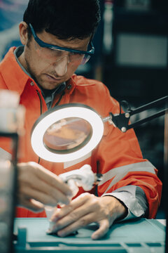 Robotics Engineer at Work: A focused engineer in safety glasses and an orange vest meticulously assembles a miniature robotic arm under the bright light of a magnifying lamp.