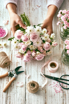 Hands are arranging a bouquet made of light pink roses and white chrysanthemums on a rough wooden table. Gardening tools are nearby