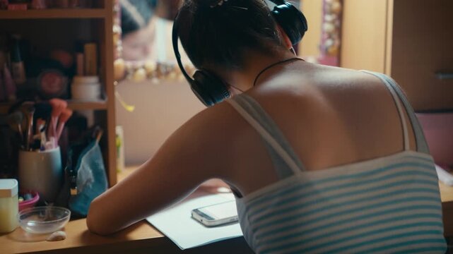 Rear view of a young female student sitting at her desk, wearing over-ear headphones and listening to music while trying to focus on textbook notes. Retro analog cinematic film look, 4k.