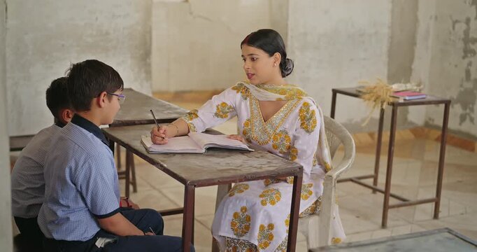 Indian young adult female sitting on bench chair boy do study learn new lesson at class room place teen child wear uniform look tutor read book talk speak share exam note day time rural area desi life