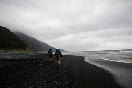 View of two hikers with backpacks walking along the black sand beach of the Lost Coast under a cloudy sky Humboldt County, California, United States.