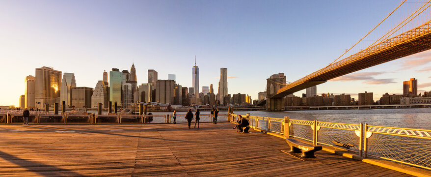 People walking and sitting on a wooden pier viewing the skyline at sunset, representing an urban travel lifestyle by the East River, Lower Manhattan, Brooklyn Bridge, New York City.