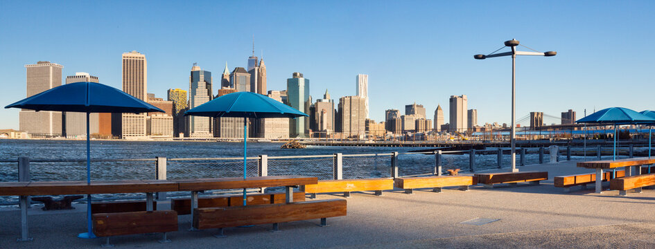Outdoor picnic tables with blue umbrellas on a waterfront promenade with skyline views, symbolizing urban tourism in New York City, Lower Manhattan, East River, Brooklyn Bridge.