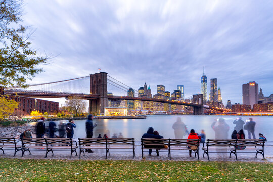 Tourists and locals enjoy a scenic urban lifestyle, relaxing on park benches while viewing the iconic Lower Manhattan skyline and Brooklyn Bridge at twilight in New York City, USA.