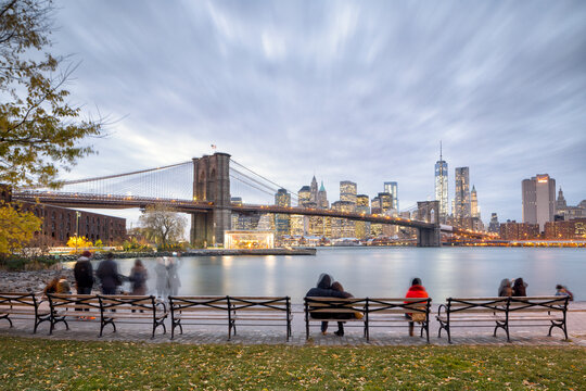 Travelers and residents relax on park benches at dusk, capturing the scenic urban lifestyle and city exploration in New York City, East River, Lower Manhattan, Brooklyn Bridge.