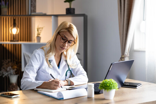 Female doctor in white coat and stethoscope writing medical notes in a notebook at her desk in a contemporary clinic office