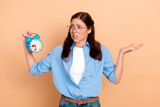 Young woman holding alarm clock and looking frustrated about time and being late, stressed about schedule and deadlines while checking the clock in a casual studio portrait