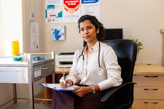 Female medical practitioner sitting beside her desk in the clinic holding a blue clipboard and a pen