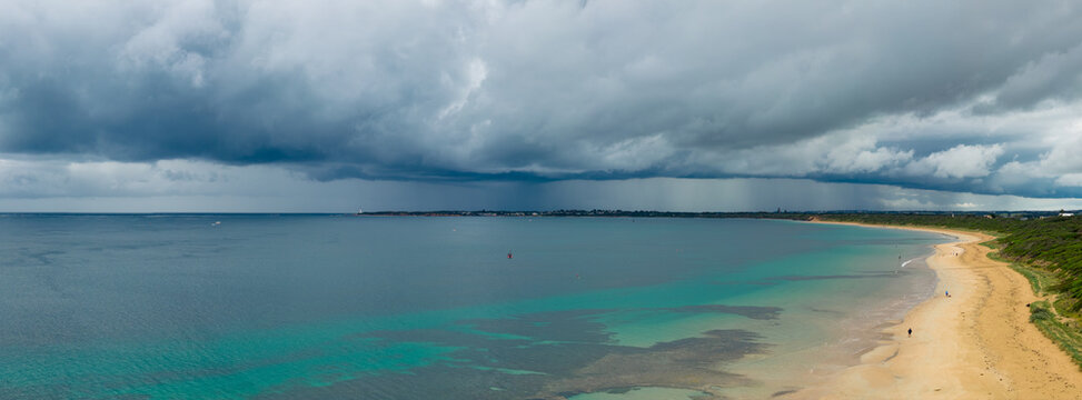 Aerial panorama of rain falling from a wide stormfront advancing over a calm bay and sandy beach