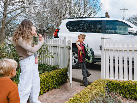 Aussie mum carrying toddler boy waving goodbye to young son near the picket fence