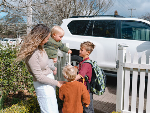 Aussie mum stands beside picket fence carrying toddler boy sending older son for school