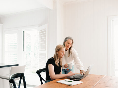 Mum leaning over teenage daughter working on her laptop on the wooden table