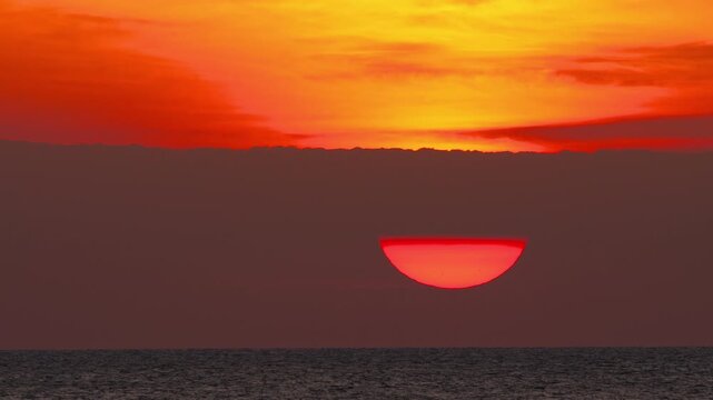 Large red sun setting behind dark clouds over calm ocean with vibrant orange sky. Telephoto close-up shot of glowing half sun disk over sea.