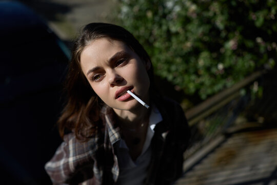 A thoughtful woman in casual attire pauses outdoors, smoke curling from a cigarette as soft sunlight highlights her features, capturing a candid lifestyle moment