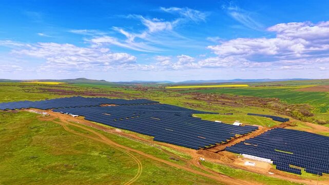Wide aerial shot of solar panel field in green landscape. High angle landscape showing a solar module array positioned within green fields under a bright blue cloudy sky.