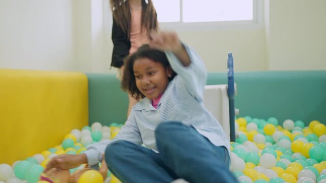 Childhood Joy in Ball Pit: A young girl of African descent smiles gleefully as she slides into a colorful ball pit, embodying pure childhood fun and innocence.