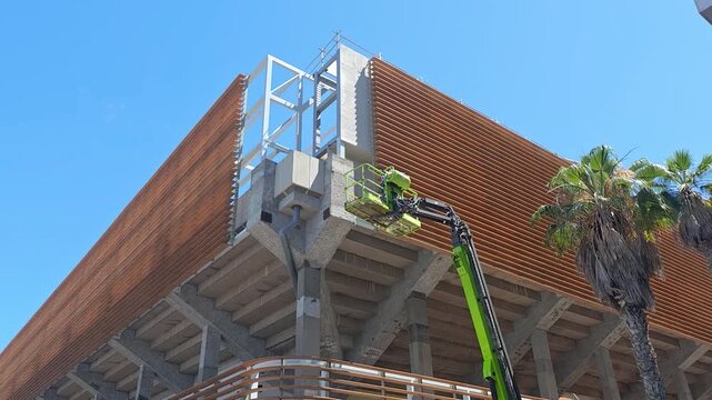 Construction Worker in Aerial Lift Basket Working on Wooden Building Facade at Construction Site during construction activities on a modern building site. Unrecognizable Worker.