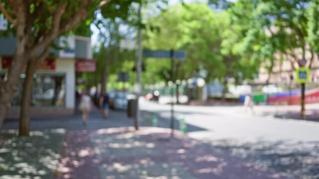 Blurred scene of people walking in a sunlit urban street with shops and green trees, creating a bokeh effect in an outdoor city environment