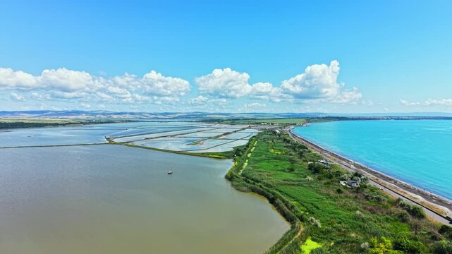 A bright day shows the salt pans of Burgas beside the Black Sea. Fields stretch along the coast with water on one side and a blue sky overhead. Clouds float in the distance.