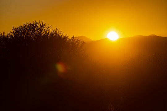 View of the sun setting over rolling hills and silhouetted bushes under a vibrant orange sky near Mazenod, Maseru, Lesotho.