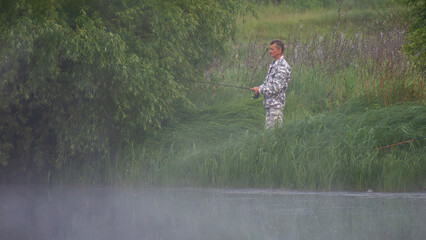 a man in camouflage clothing fishing by a misty lake or river, surrounded by lush green trees © Александр Арендарь