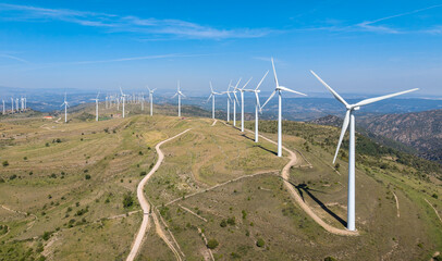 Aerial view of a large wind farm stretching along a mountain ridge in the Maestrat region, Spain, with turbines lining a dirt road under a clear blue sky © IgorsPerspective