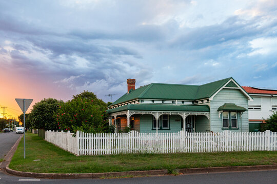Dramatic sunset sky over green weatherboard house behind white picket fence