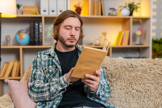 Young man on home sofa reads a few lines in book then turns page and pauses in thought. Adult guy on home sofa looks ahead for a moment while reflecting on plot meaning and ideas from reading quietly.