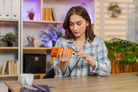 Young woman on home table tries to connect charger to smartphone but cable fails during work. Adult girl on home table looks worried and puzzled by smartphone problem and charging malfunction at desk.