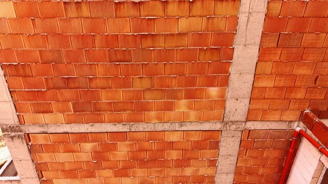 Close up view of red brick wall under construction background. Factual detailed view of an unfinished hollow red ceramic brick wall within a concrete structural frame with pipes.