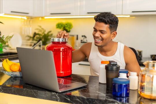 Indian man explains sports nutrition and workout supplements on laptop during meal planning at home. Arabian guy shares training advice and protein diet tips with focused face in home kitchen indoors.