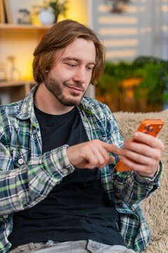 Young man on home sofa types quick smartphone message then stops to reread words with focused smile. Adult guy on home sofa presses send with calm satisfaction after checking text and finishing chat.