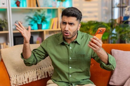 Indian man on home sofa speaks on smartphone speaker call with stop gesture and upset look in tense argument. Arabian guy on home sofa reacts with stress and anger in loud phone dispute indoors alone