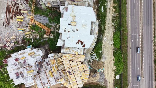 Workers are present at a large construction site next to a busy road. Vehicles drive by while materials are scattered across the area. The project shows signs of heavy activity during daytime.