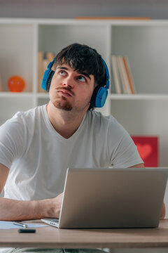 Young man at home taking an online course with headphones and a laptop on his desk, studying and working with a thoughtful expression
