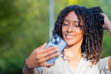 Obraz premium young latin woman laughing while taking a selfie with smartphone outdoors