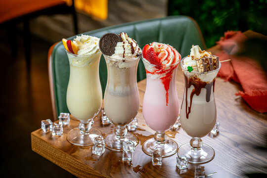 Different milkshakes line up on table. Four milkshakes sit in tall glasses on a wooden table. Each shake is topped with cream and fruits.