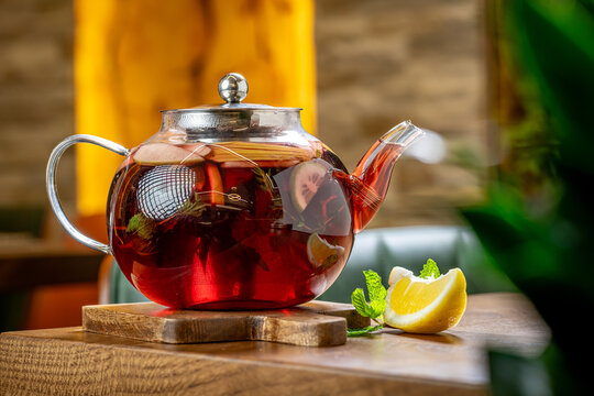 Brewing tea in a glass pot with lemon. A glass teapot sits on a wooden board with tea inside. Lemon slices are next to the pot on the table.