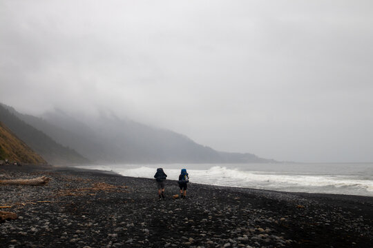 View of two hikers with backpacks walking along the dark pebbly shore of the Lost Coast under a foggy sky with misty mountains in Humboldt County, California, United States.