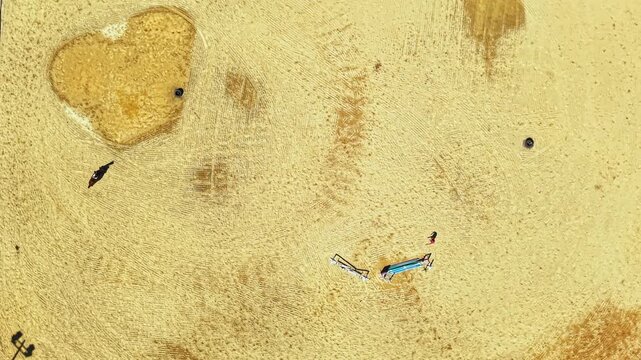 People are gathered on a sandy area. Two boats are placed nearby. A heart shape is formed in the sand. The scene is bright and sunny near the water.