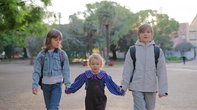 Three siblings walk hand in hand through urban park with palm trees and buildings visible in background. Older children escort younger sister across paved plaza during golden hour light.