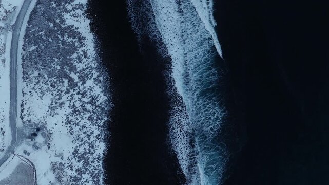 Top down aerial of dark Arctic coast bordered by snowy beach, drone establishing background, waves crash with whitewash on rocks