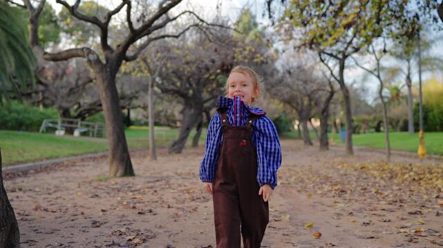 Adorable young girl with curly blonde hair dressed in blue gingham shirt and brown corduroy overalls smiles while walking toward camera through scenic autumn park with mature trees and scattered.