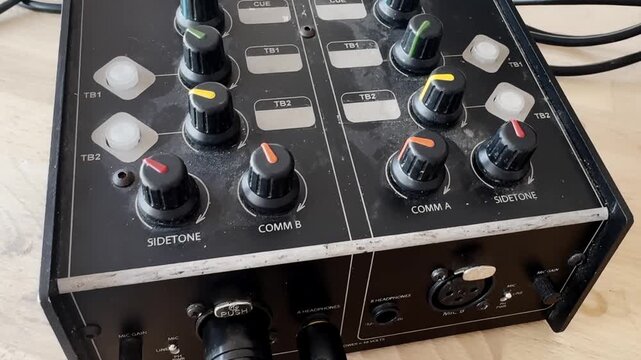 Close-up of an audio intercom control unit with colorful knobs, talkback buttons, sidetone controls and XLR connectors on a wooden table.