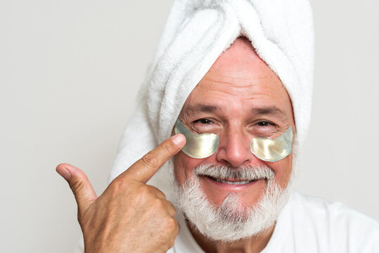 Happy senior man with gray beard wearing towel on head and applying eye patches for skincare