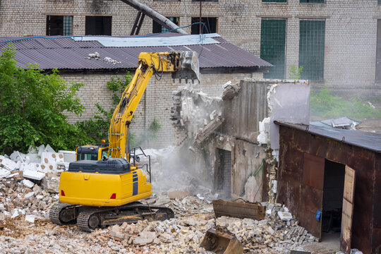View of a yellow excavator demolishing a brick wall with dust and debris at a construction site in Vilnius, Vilnius County, Lithuania.