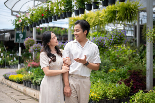 Asian man and woman couple linking arms chatting together beside colorful plants in nursery garden.